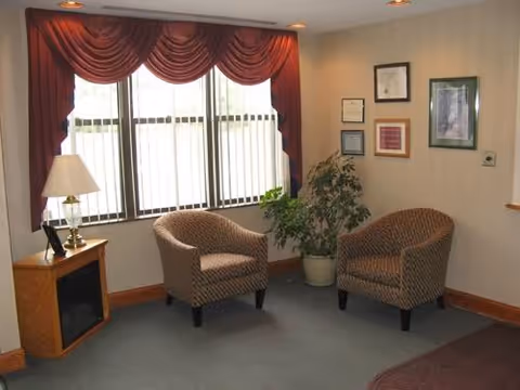 A small sitting area with two patterned armchairs, a potted plant, a wooden side table with a lamp, and a window with vertical blinds and red valance curtains. The walls have framed pictures and certificates.