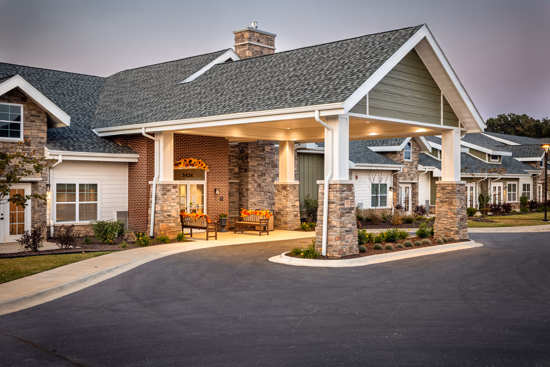 Exterior view of a senior living facility named Ciel of Fayetteville during dusk. The building features a covered entrance with stone pillars and a peaked roof. There are benches with fall-themed decorations near the entrance door, which has the number 3434 above it. The building exterior combines brick, stone, and siding with multiple windows and a well-maintained driveway and landscaping.