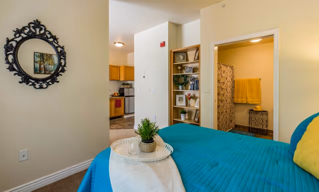 Bedroom interior with a teal bedspread, decorative round mirror on the wall, and open doorways showing a kitchen and a bathroom.