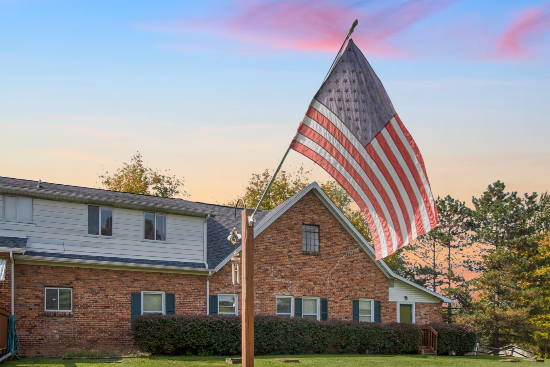 Exterior view of a brick and siding building with multiple windows and green shutters, surrounded by trees and bushes. An American flag is prominently displayed on a flagpole in the foreground against a colorful sky at sunset.