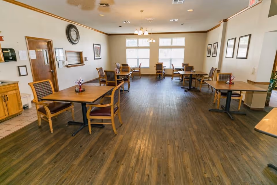 A dining room in Meadow Creek Senior Living featuring several wooden tables with chairs arranged around them. The room has large windows letting in natural light, framed pictures on the walls, a clock above a service window, and a wooden floor.