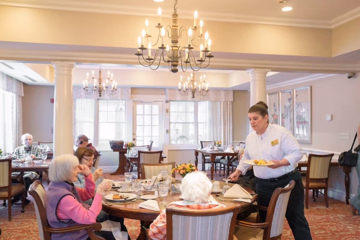 A dining room in Orchard Hill at Sudbury with elderly residents seated around a table enjoying a meal. A staff member is serving food to one of the residents. The room is well-lit with chandeliers, large windows, and decorated with flowers on the tables.