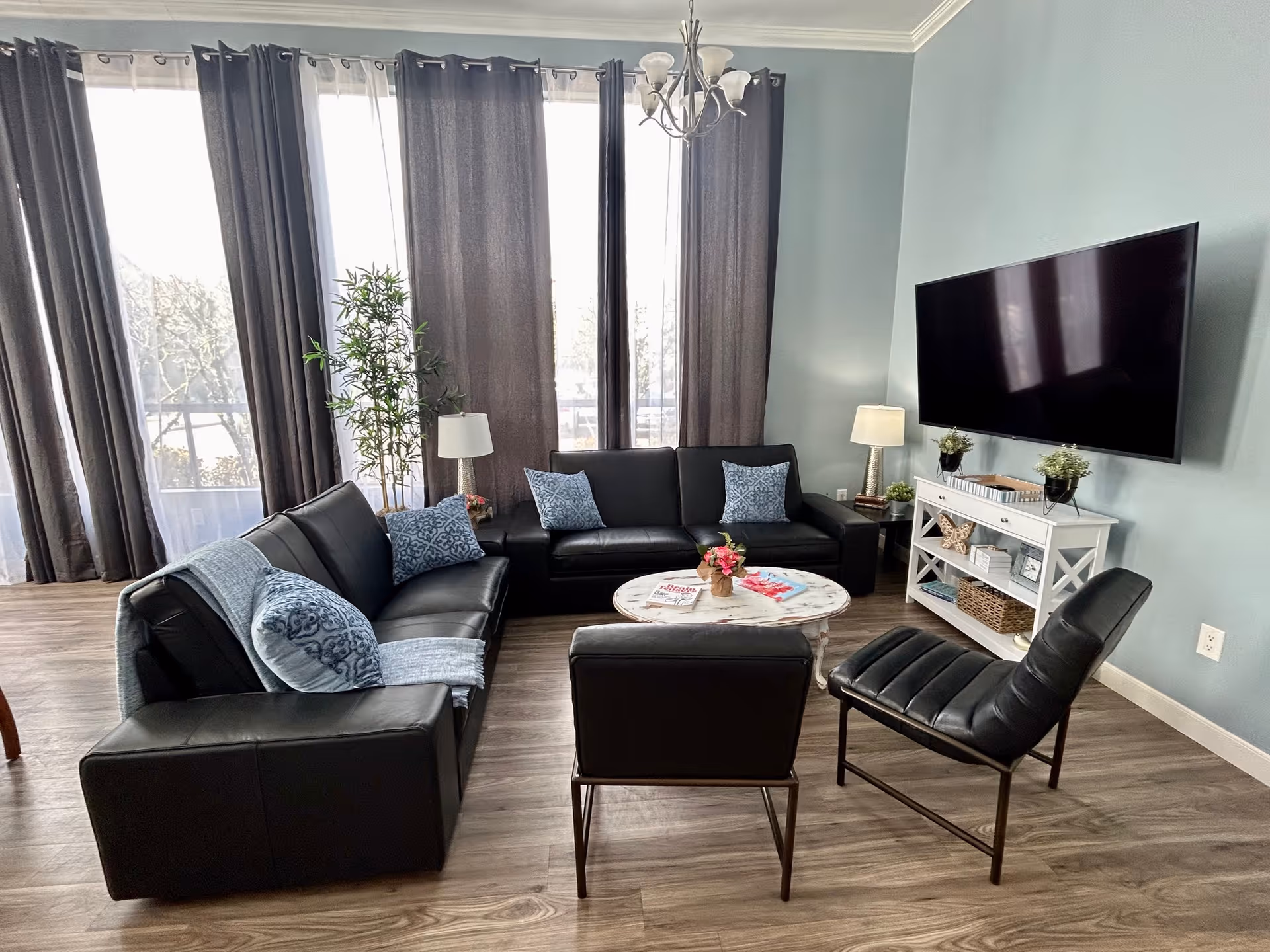 A modern living room with two black leather sofas, two black leather chairs, and a round white coffee table with a flower arrangement and magazines on it. The room has large windows with dark curtains, a light blue wall with a mounted flat-screen TV, a white console table with decorative items, two table lamps, and a tall plant near the window. The floor is wood with a light finish.