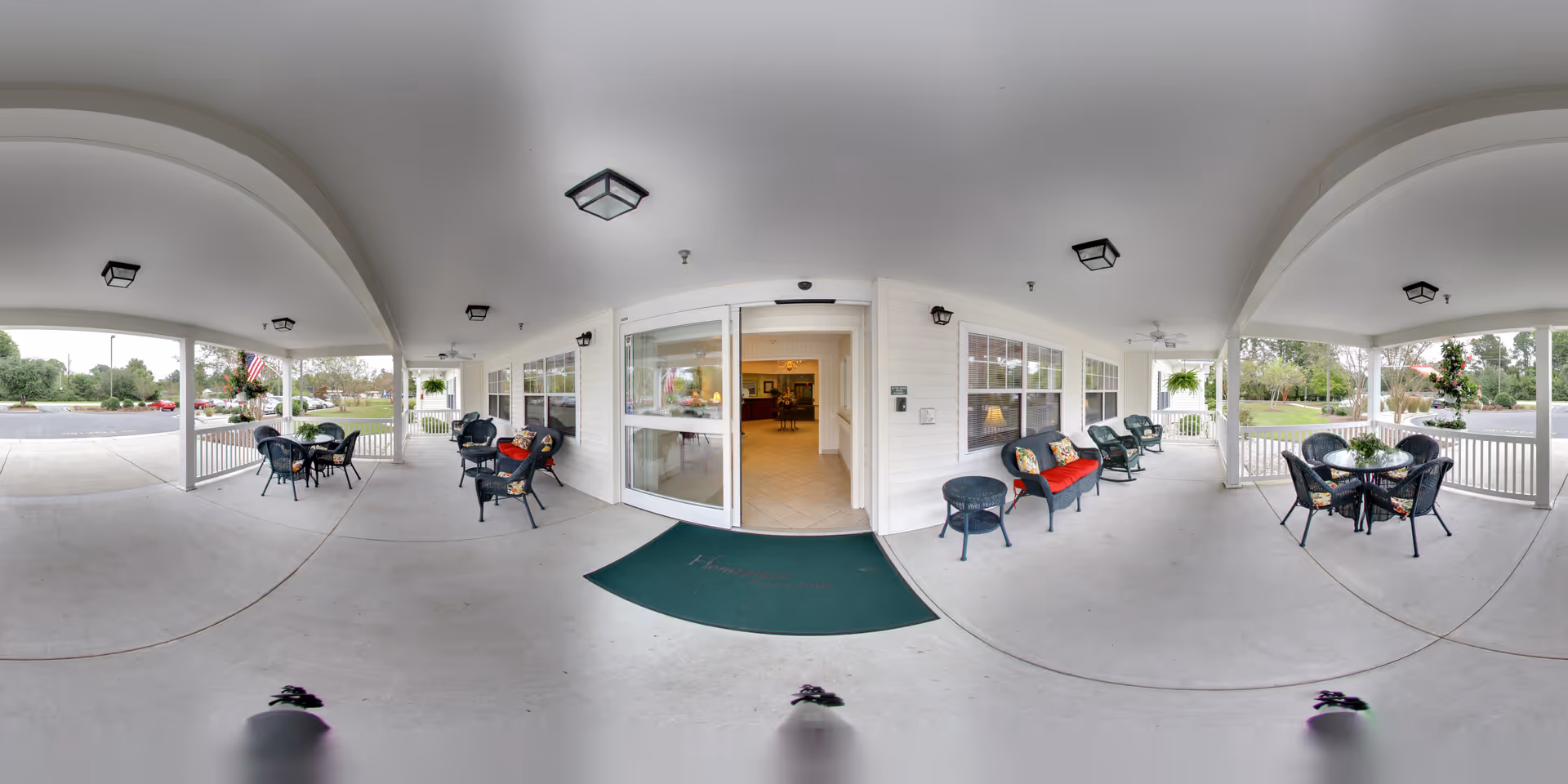 Covered outdoor patio area at Home Place of New Bern with multiple seating arrangements including black chairs and tables. The patio has a white ceiling with mounted lights and ceiling fans. There are potted plants and decorative cushions on some chairs. The entrance to the building features sliding glass doors with a green welcome mat in front. The surrounding area includes a parking lot and greenery.
