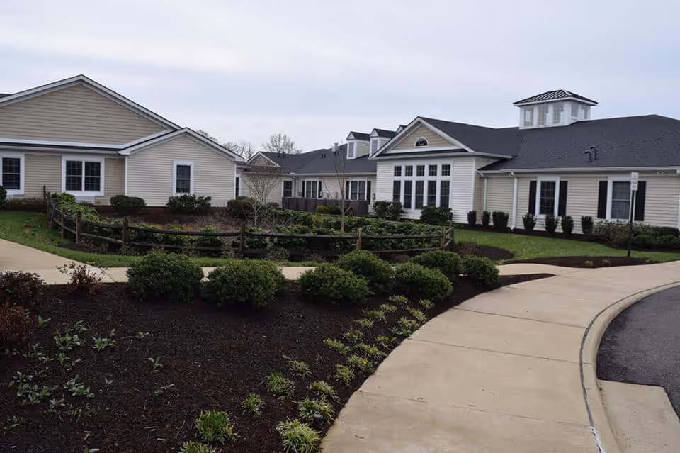 Exterior view of a senior living facility with beige siding, multiple windows, and a black roof. The foreground features a curved concrete walkway bordered by landscaped garden beds with bushes and small plants. A wooden fence encloses part of the garden area.