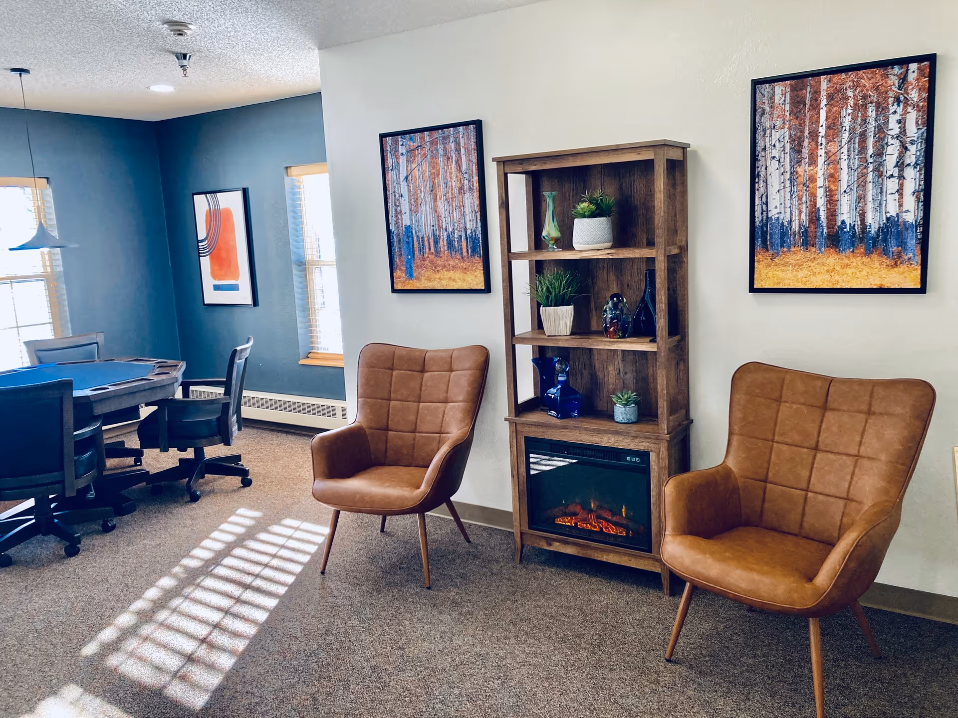 A cozy interior room featuring two brown leather armchairs with a wooden shelving unit between them. The shelving unit holds decorative items and has an electric fireplace at the bottom. The walls are adorned with framed artwork depicting autumn trees. In the background, there is a table with several chairs around it, set against a blue accent wall with windows letting in natural light.