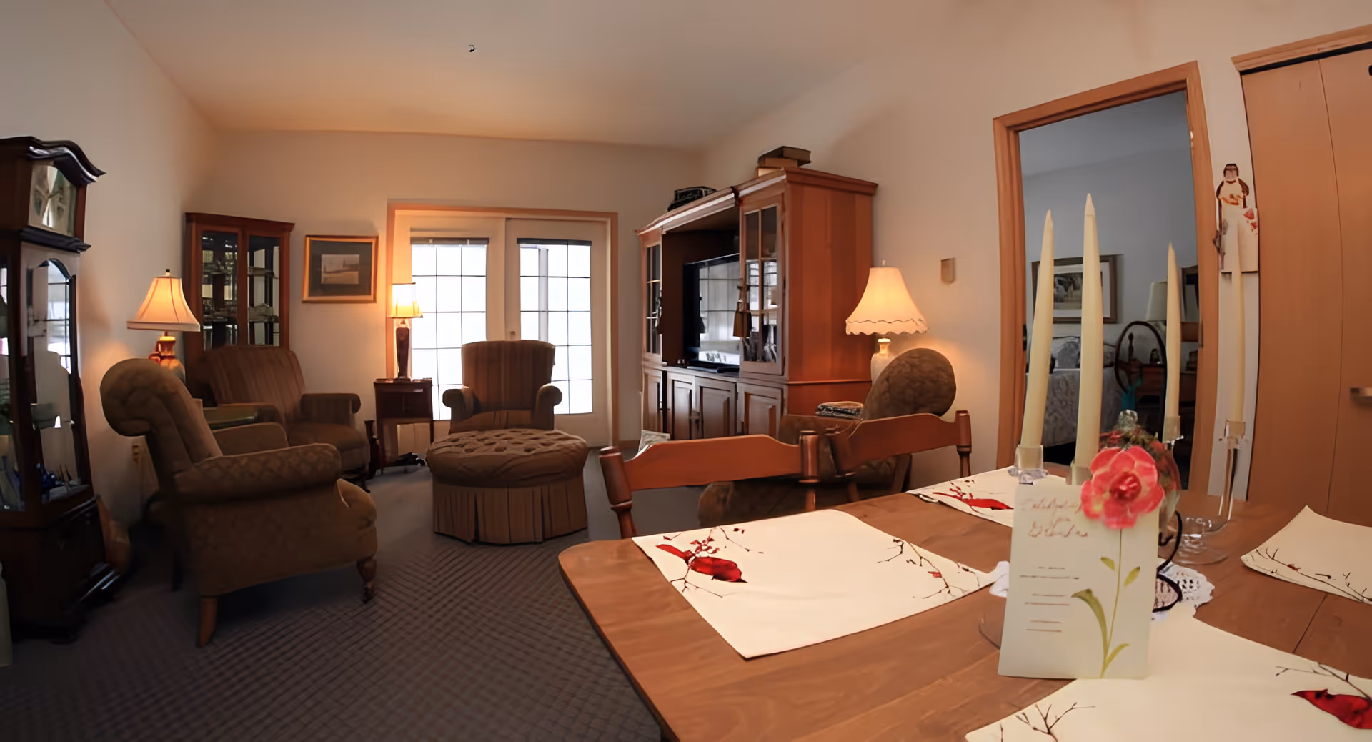 A cozy living room and dining area with upholstered armchairs, a wooden entertainment center with a TV, a wooden dining table set with placemats and tall candles, and soft lighting from table lamps. The room has carpeted flooring and a door leading to another room.