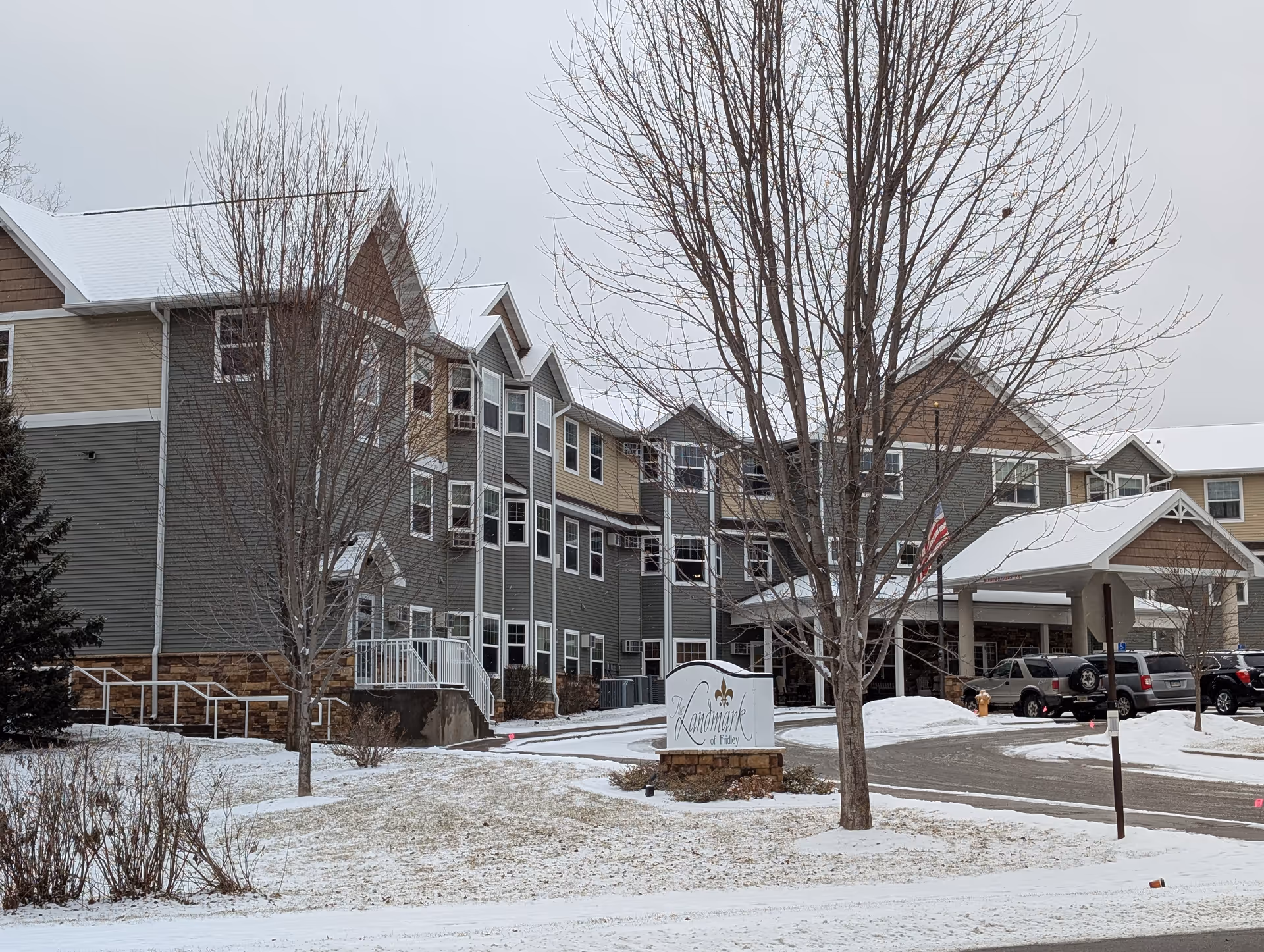 Exterior view of The Landmark of Fridley senior living facility during winter with snow on the ground and roof. The building is multi-story with gray and beige siding, multiple windows, and a covered entrance with parked cars nearby. Leafless trees and a sign with the facility name are visible in the foreground.