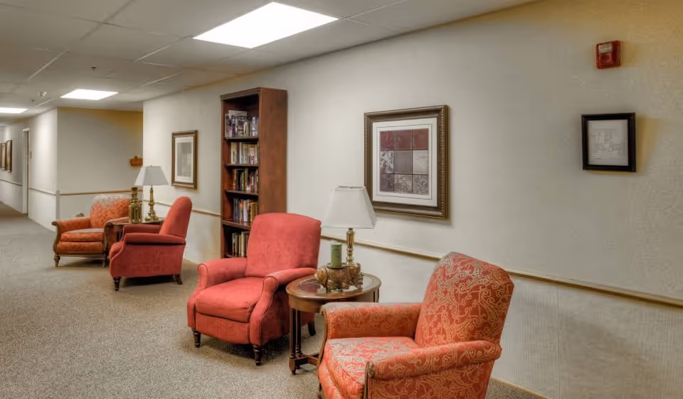 A hallway in a senior living facility with several upholstered armchairs in red and orange patterns arranged along the wall. There are small side tables with lamps and decorative items between the chairs. A tall bookshelf filled with books is also visible. The walls are decorated with framed artwork and the floor is carpeted.