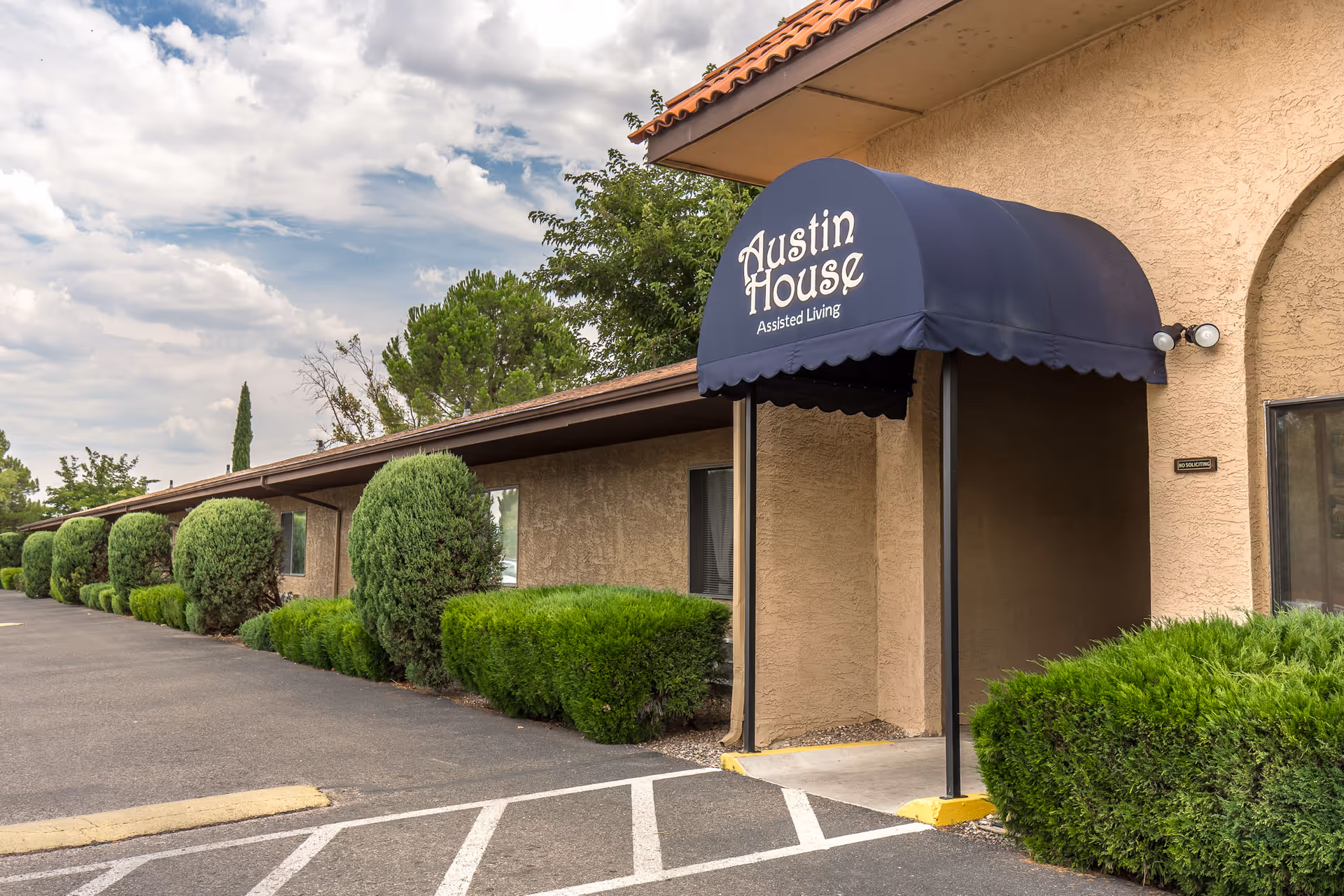 Exterior view of a single-story assisted living facility with a beige stucco exterior and a blue awning that reads 'Austin House Assisted Living'. The building is surrounded by neatly trimmed bushes and trees, with a paved parking area in front under a partly cloudy sky.
