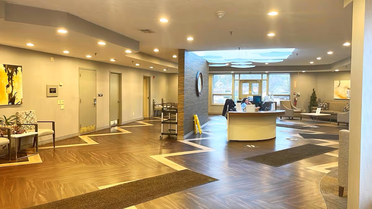 Interior view of a senior living facility lobby with a reception desk where two staff members are seated. The space features wood-patterned flooring with geometric designs, recessed ceiling lights, and modern furniture including chairs and small tables. Large windows in the background allow natural light to enter, and there is a decorative wall with a clock behind the reception desk.