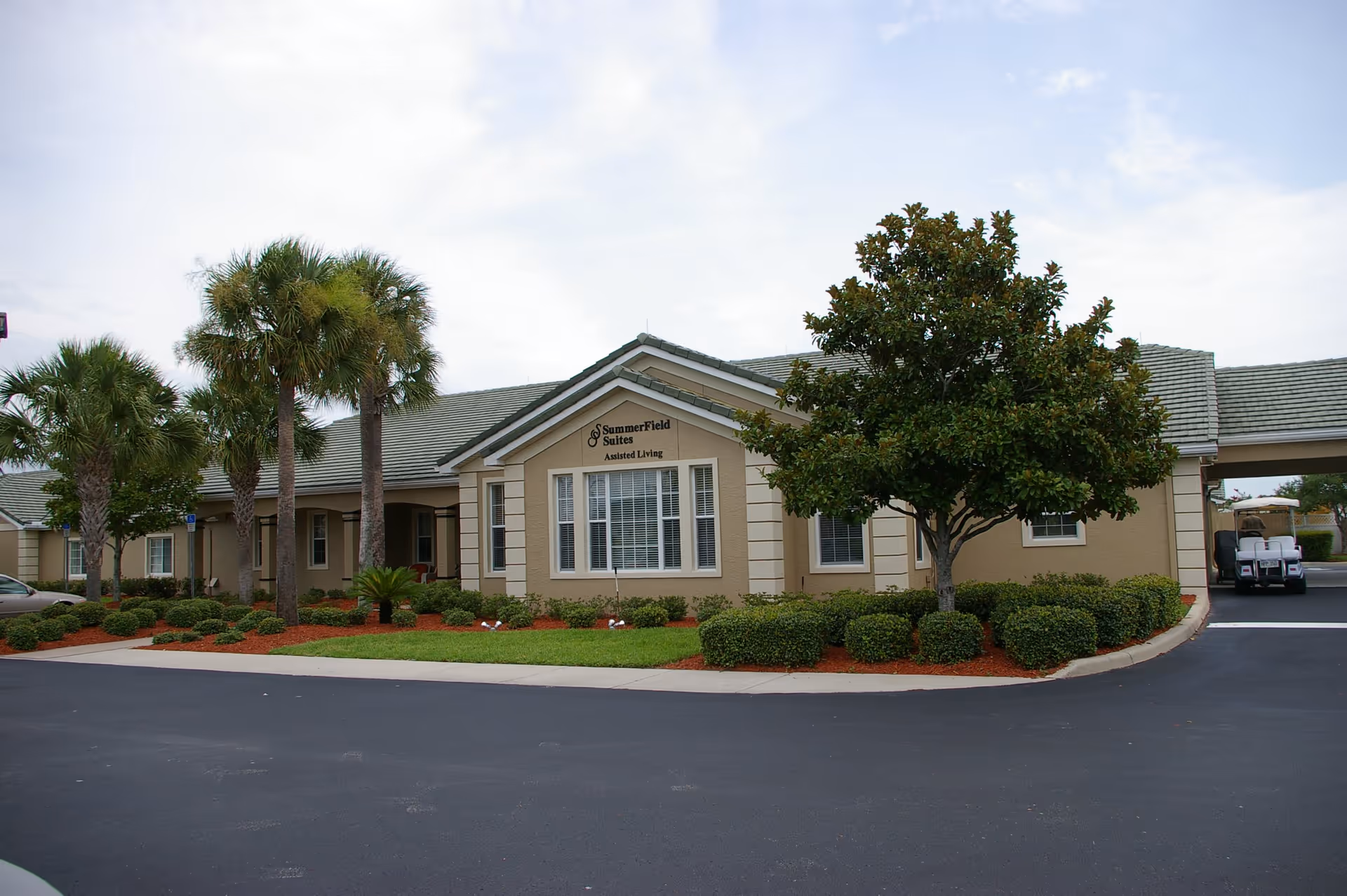 Exterior view of Summerfield Suites Assisted Living facility showing a beige building with green roof, surrounded by palm trees, bushes, and a well-maintained lawn under a partly cloudy sky.