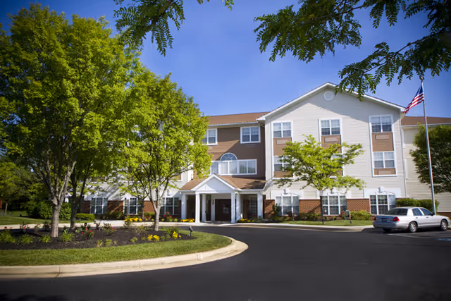 Exterior view of a three-story senior living facility building with a driveway and landscaped greenery including trees and bushes. An American flag is visible on a flagpole near the right side of the building.