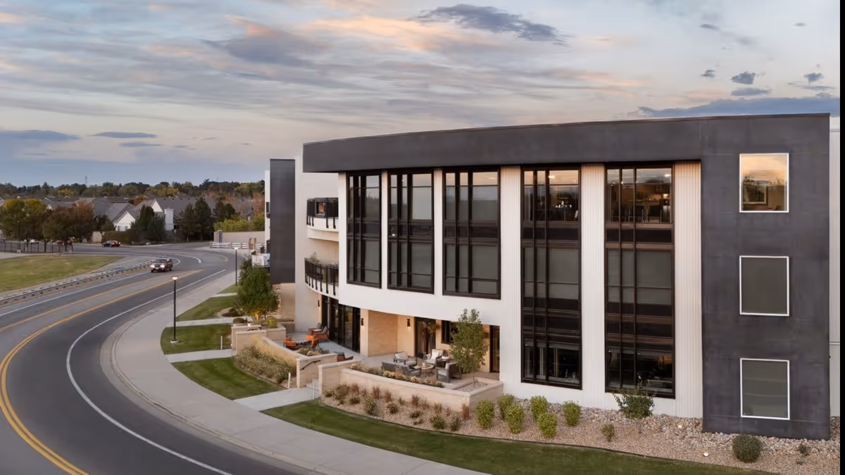 Exterior view of a modern, multi-story building with large windows and outdoor seating areas, situated along a curved road with a few cars and street lamps, under a partly cloudy sky during dusk.