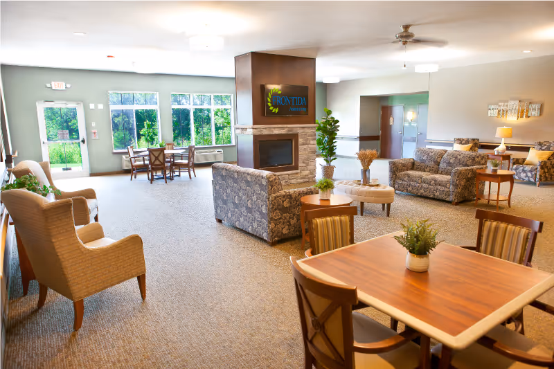 A spacious and well-lit assisted living facility common area with multiple seating arrangements including armchairs, sofas, and tables with chairs. There is a fireplace in the center with a sign above it that reads 'Frontida Assisted Living'. Large windows let in natural light and offer a view of greenery outside. The room has neutral-colored walls and carpeted floors, with plants and decorative items placed around.