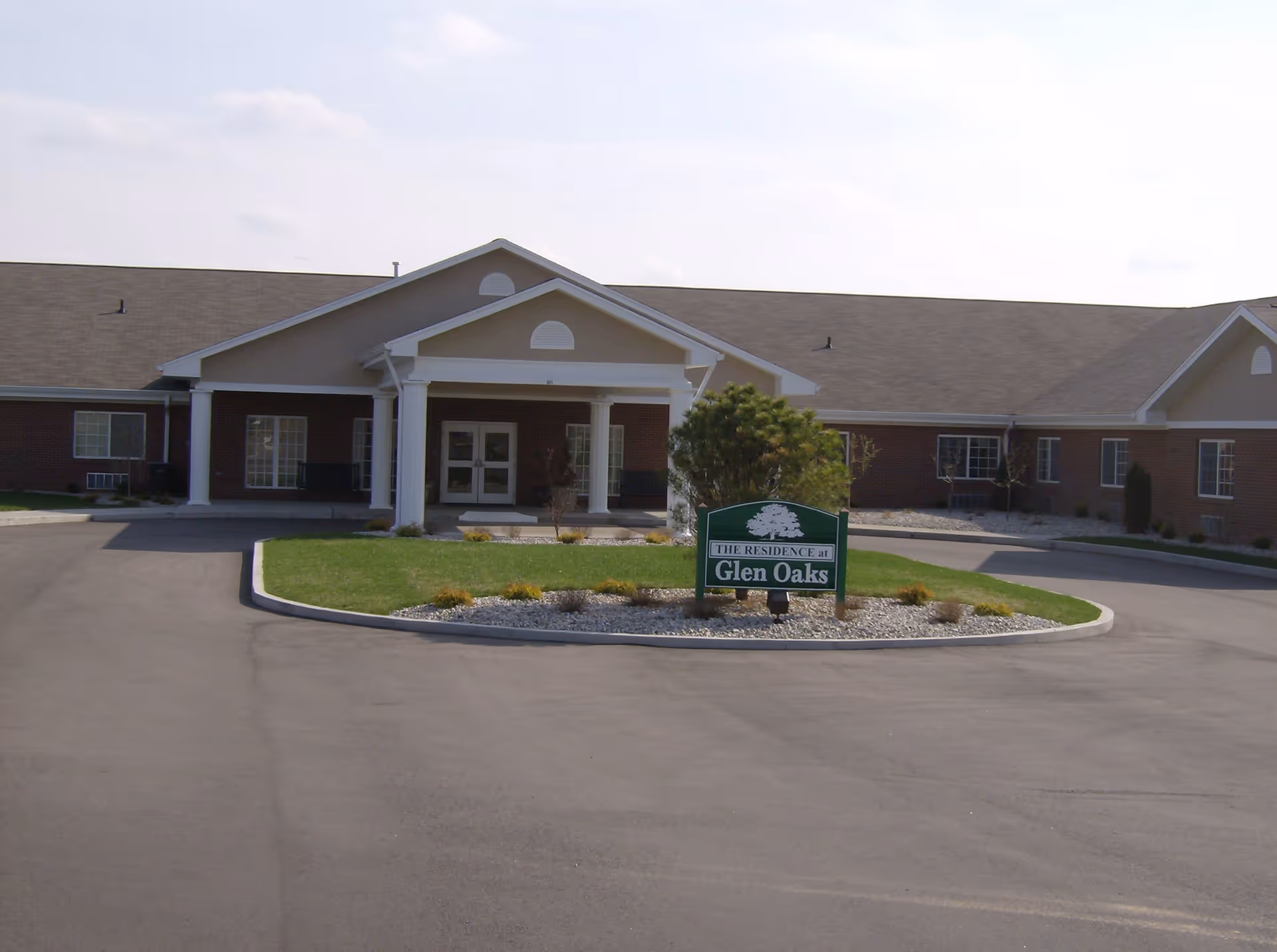 Front exterior view of Glen Oaks Health Campus building with a covered entrance supported by white columns, a circular driveway, and a green sign in the center of the landscaped area that reads 'The Residence at Glen Oaks'.