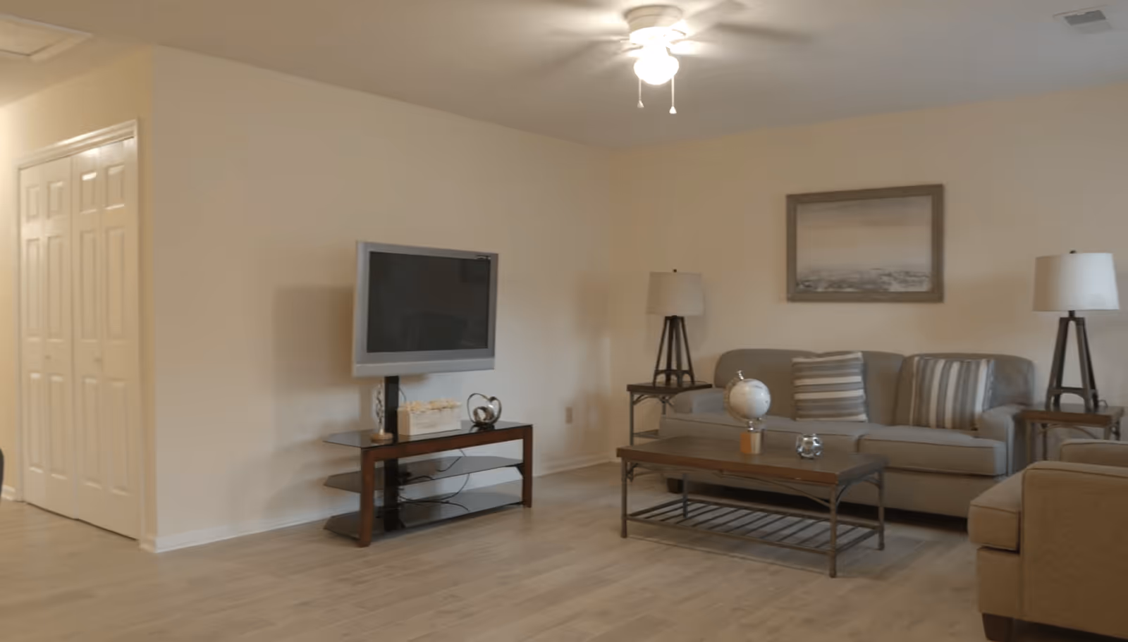 A living room with a beige couch, two side tables each with a lamp, a coffee table with a decorative globe and glass item, a wall-mounted flat-screen TV on a wooden stand, light-colored wooden flooring, and a ceiling fan with a light. There is a framed picture hanging on the wall above the couch.