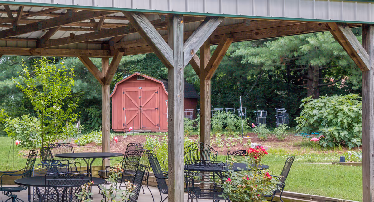 Outdoor covered patio area with black metal tables and chairs, surrounded by greenery and a garden with a red wooden shed in the background.