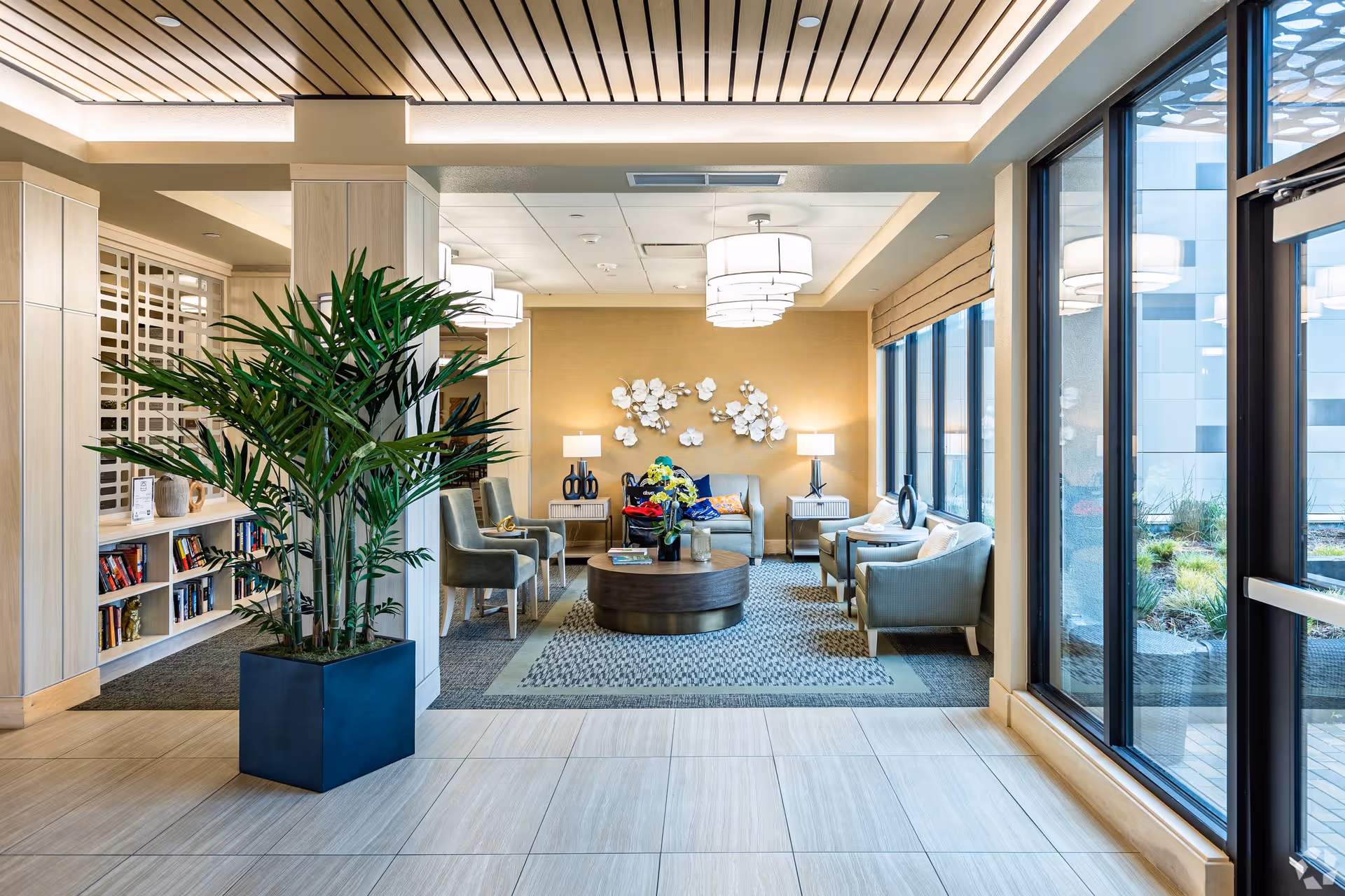 A bright and modern lounge area with large windows on the right letting in natural light. The room features a seating arrangement with armchairs and a sofa around a round coffee table on a patterned rug. There are two side tables with lamps and decorative wall art above the sofa. A large potted plant is positioned near the entrance, and built-in shelves with books are visible on the left side.