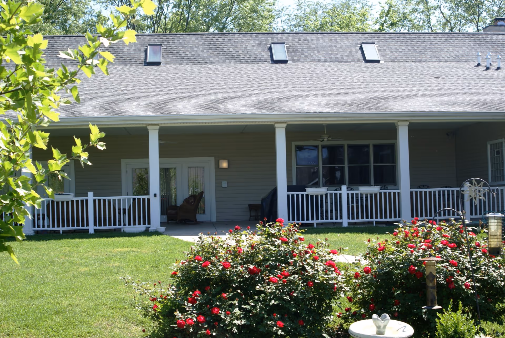 View of the back porch of a single-story building with a gray shingled roof and white railing. The porch has outdoor furniture including chairs and a table. In the foreground, there is a well-maintained green lawn with blooming red rose bushes and garden decorations. Trees with green leaves are visible in the background.