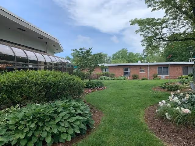 A well-maintained outdoor garden area with green grass, bushes, and flowering plants. There is a brick building in the background with windows and an air conditioning unit. The sky is partly cloudy with blue patches visible.
