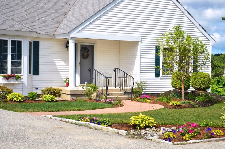 Front exterior view of a white single-story house with a small porch, black railings, a door with a wreath, green shutters on the windows, and a well-maintained garden with colorful flowers and shrubs.