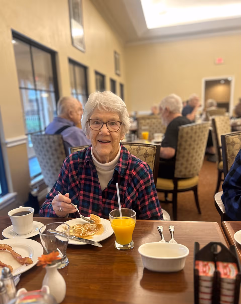 An elderly woman with white hair and glasses is sitting at a dining table in a senior living facility, eating pancakes with syrup. On the table, there is a glass of orange juice, a cup of coffee, a plate with bacon, and some utensils. Other elderly people are seated at tables in the background in a well-lit dining room with large windows.