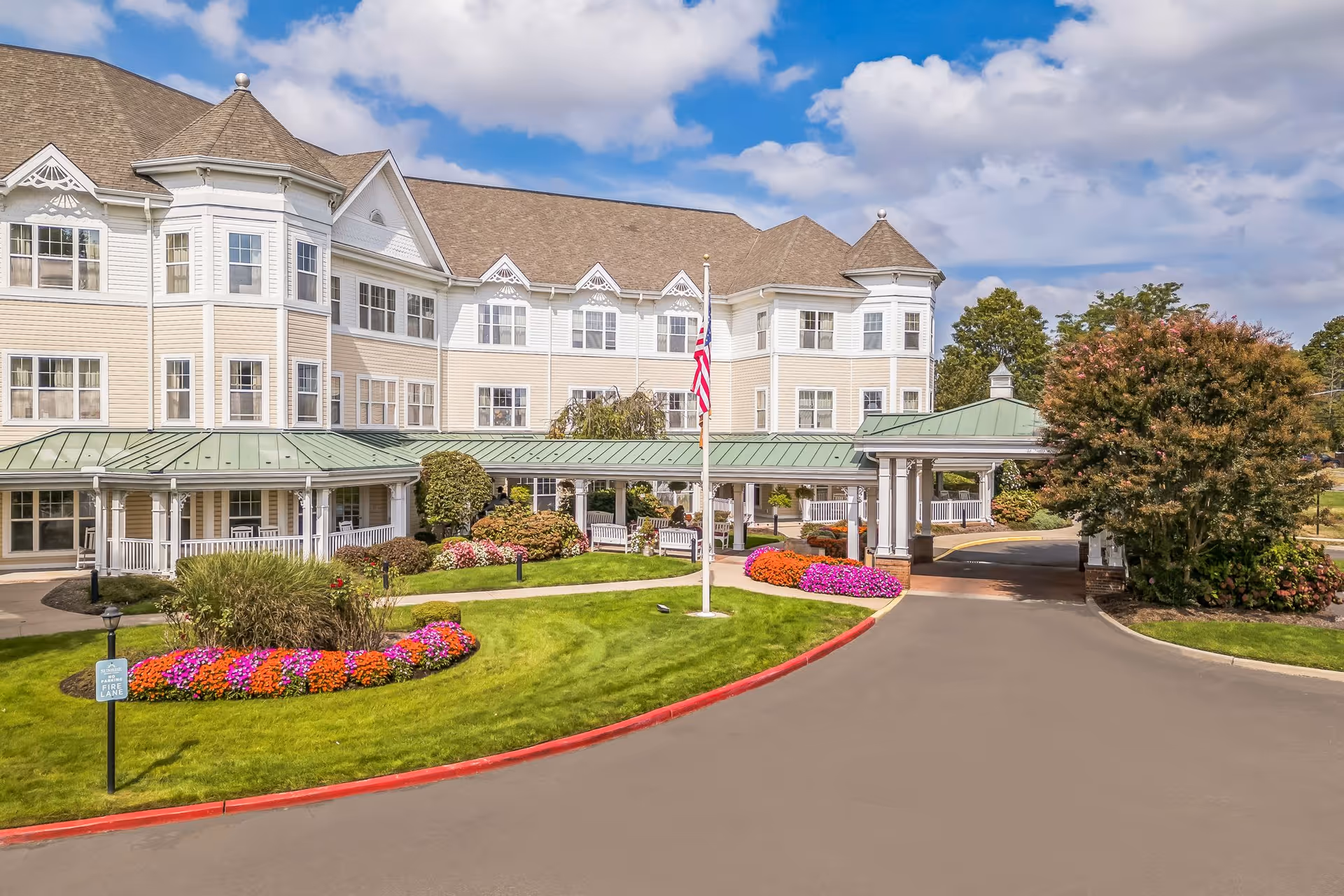 Exterior view of a large senior living facility building with beige siding and white trim, featuring multiple windows and a green metal roof over the entrance. The driveway curves around a landscaped area with colorful flowers and a flagpole with an American flag. The sky is partly cloudy.