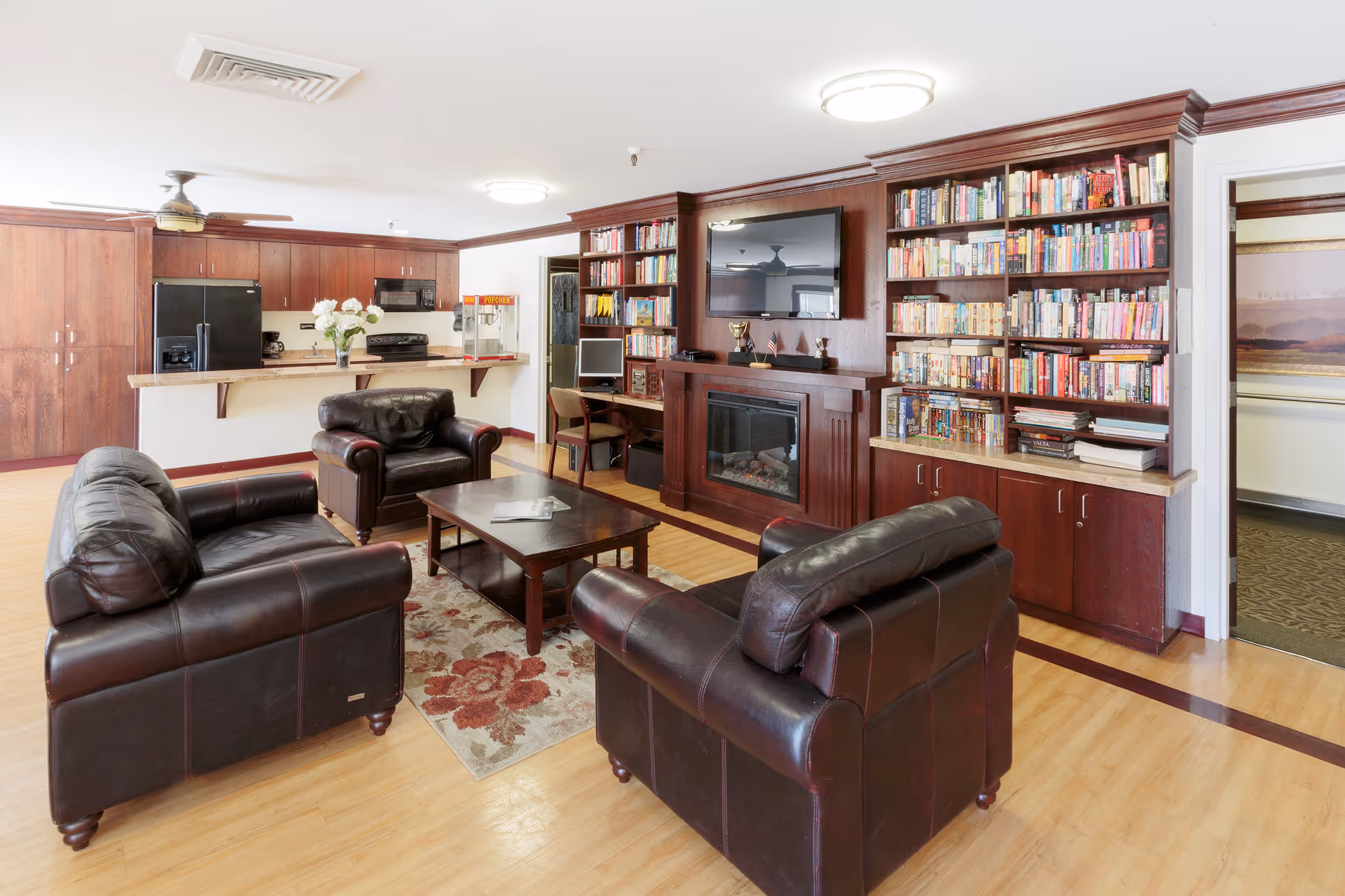 A cozy living room area in a senior living facility featuring three dark brown leather armchairs arranged around a wooden coffee table on a floral rug. Behind the seating area is a built-in wooden bookshelf filled with books and a flat-screen TV mounted above a fireplace. To the left, there is a kitchen area with wooden cabinets, a black refrigerator, a stove, and a popcorn machine on the counter. The room has light wood flooring and white walls with wooden trim.