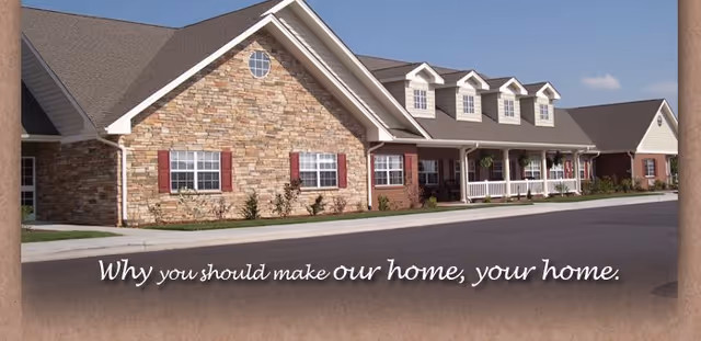 Exterior view of a single-story senior living facility building with stone and brick facade, multiple windows with red shutters, a covered porch with white railings, and a paved driveway in front.