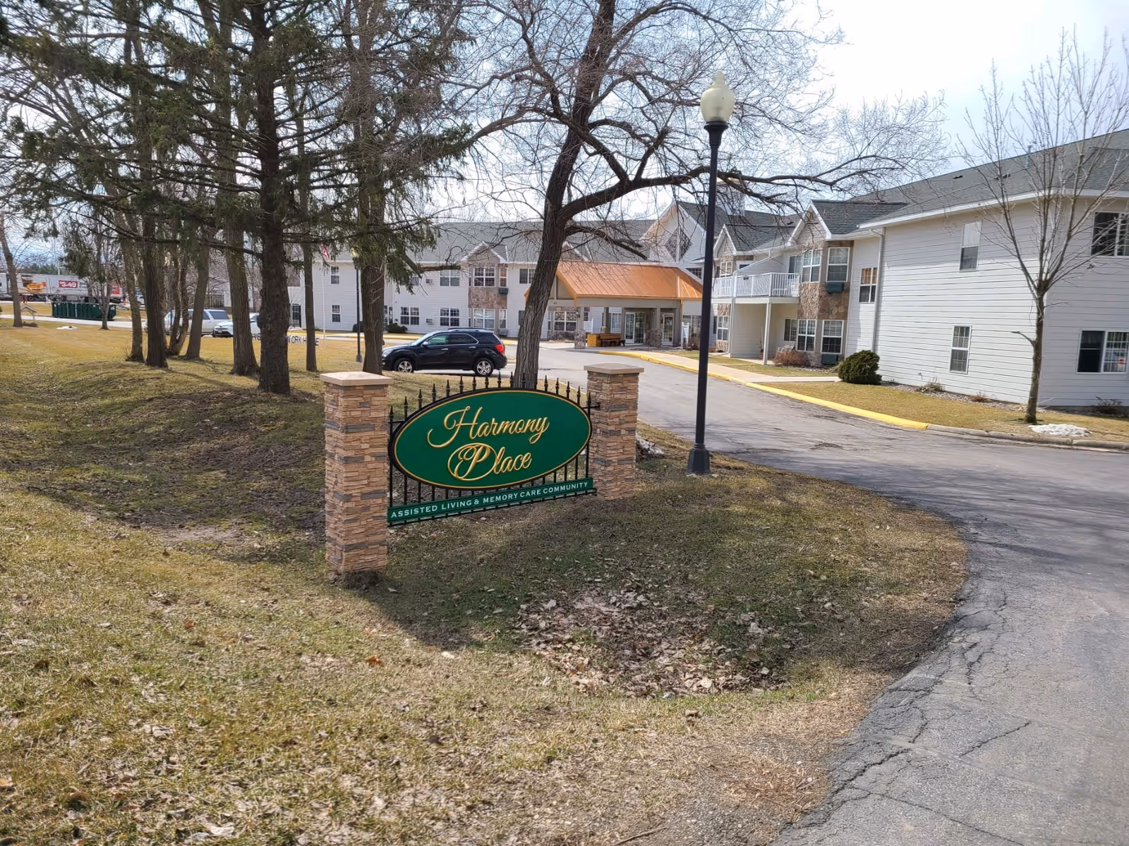 Exterior view of Harmony Place assisted living and memory care community showing the entrance driveway, a green and gold sign with the facility name, trees, and multi-story residential buildings in the background.