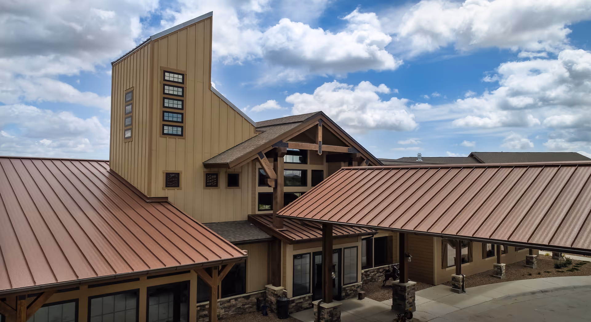Exterior view of a senior living facility building with a modern rustic design featuring tan siding, stone accents, and a reddish-brown metal roof under a partly cloudy blue sky.