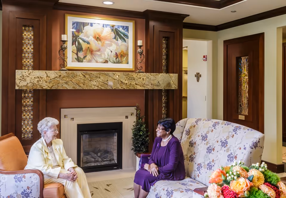 Two elderly women sitting and conversing in a cozy living room area with a fireplace, floral artwork above the mantle, and floral-patterned furniture. There is a bouquet of colorful flowers on a table in the foreground.
