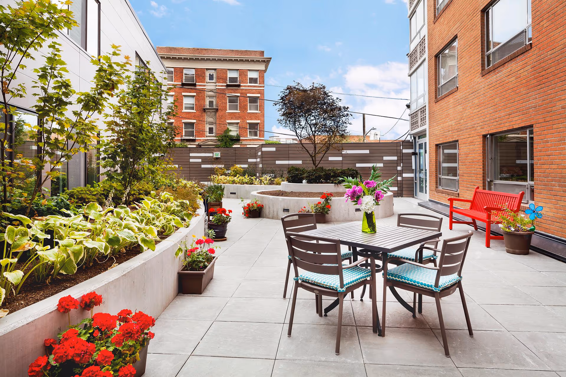Outdoor patio area at Bayview Retirement Community featuring a table with four chairs, a vase with flowers on the table, a red bench, various potted plants, and greenery along the side. The area is surrounded by buildings and a clear blue sky is visible.