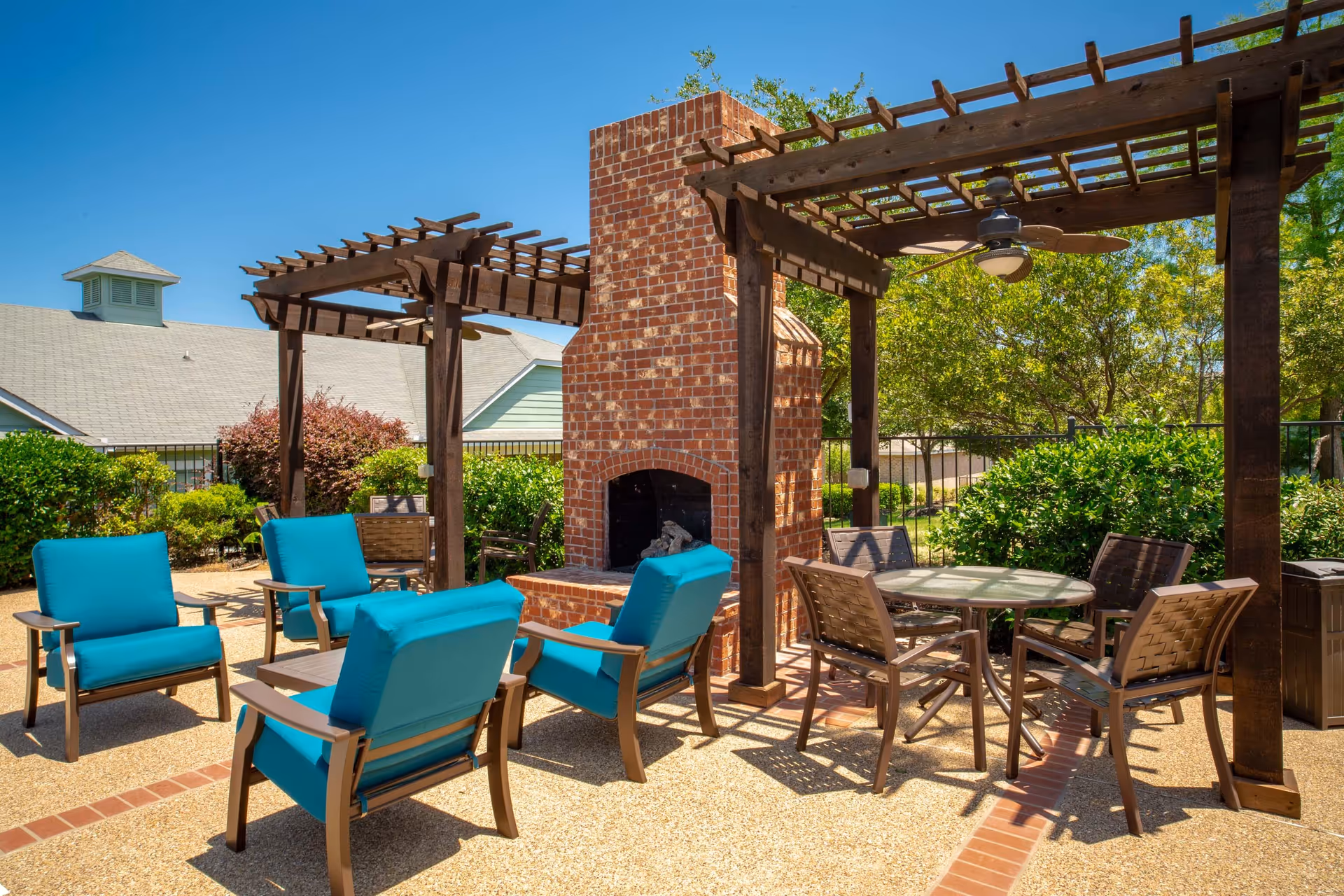 Outdoor patio area with a brick fireplace, wooden pergolas, and seating arrangements including blue cushioned chairs and a round table with four chairs, surrounded by greenery and under a clear blue sky.