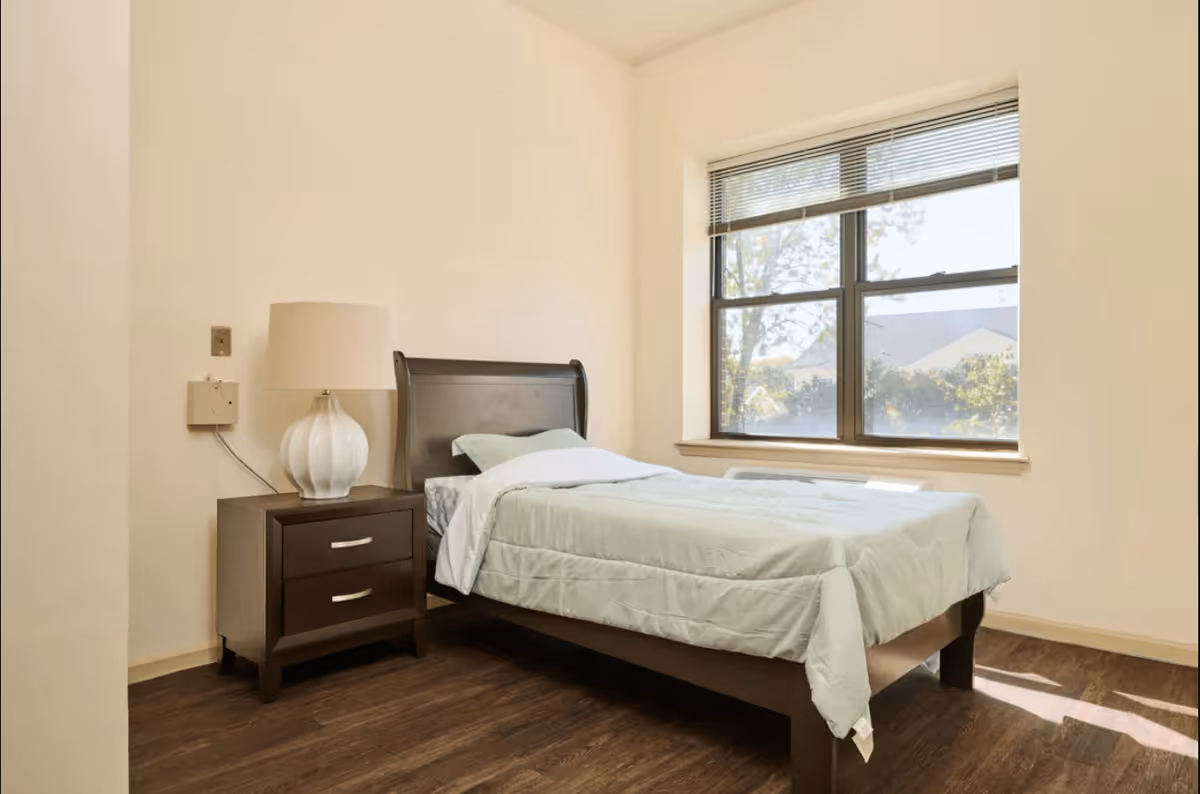 A simple bedroom with a single bed covered with light blue bedding, a dark wooden headboard, a matching nightstand with two drawers, and a white table lamp. There is a large window with blinds letting in natural light, and the floor is dark wood.