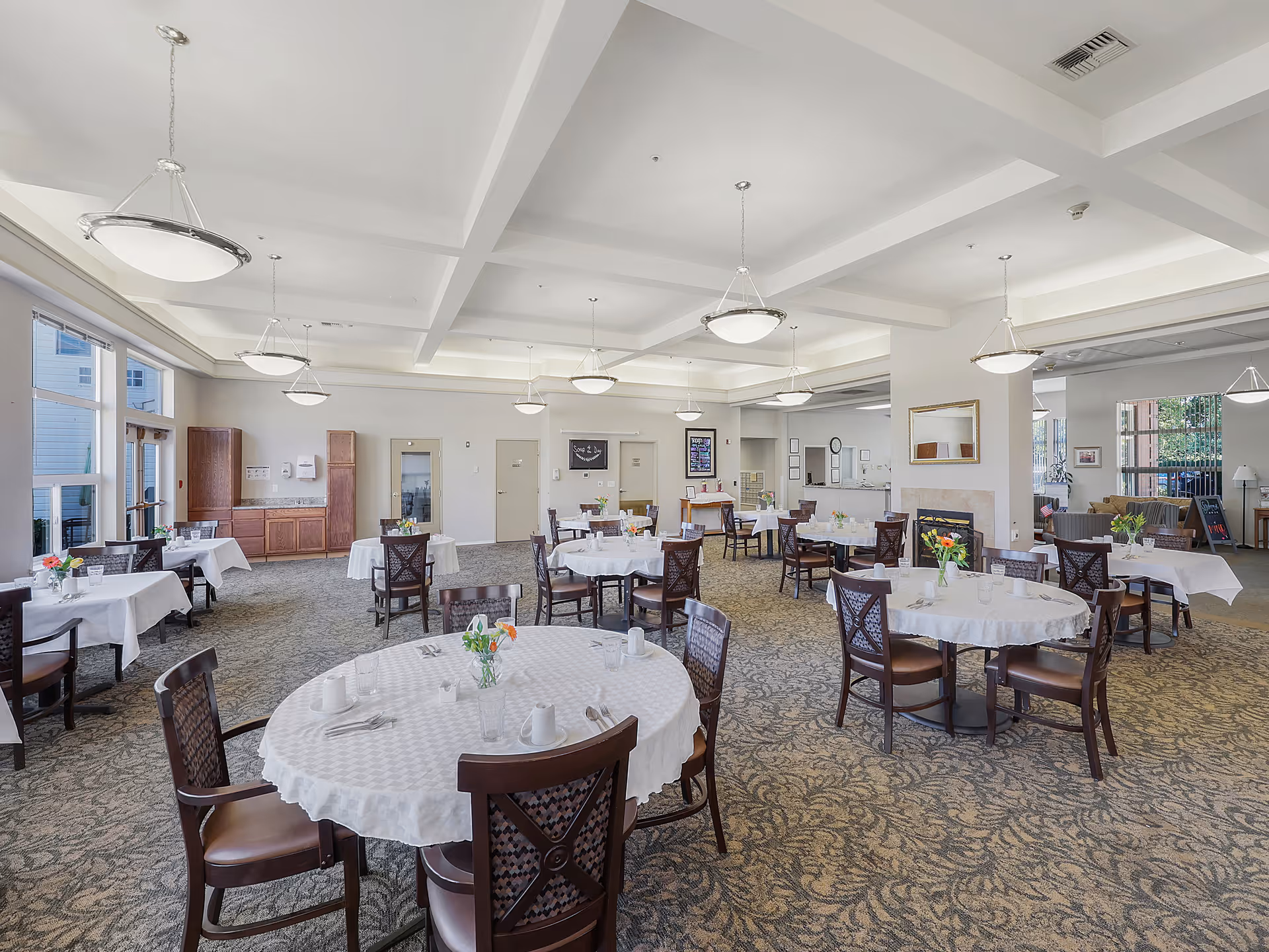 A spacious dining room with multiple round tables covered with white tablecloths, each set with cups, plates, and small flower arrangements. The room has large windows letting in natural light, modern ceiling lights, and a carpeted floor with a patterned design. There is a fireplace on the right side and a seating area visible in the background.