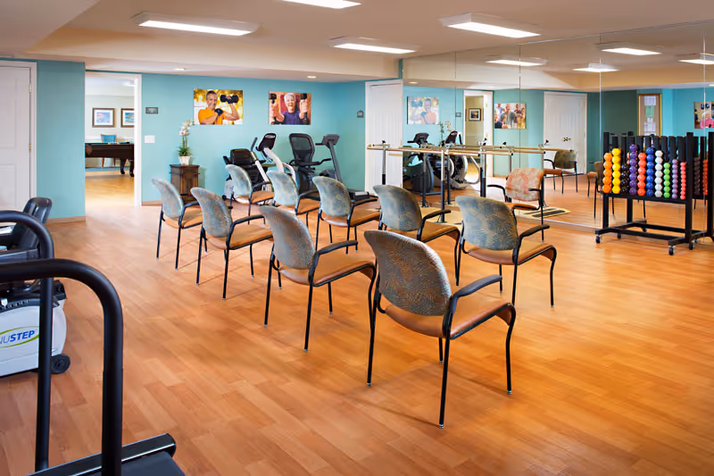 A senior living facility exercise room with wooden flooring, several chairs arranged in rows, exercise equipment including stationary bikes and a rack of colorful dumbbells. The walls are painted light blue and one wall is covered with large mirrors. There are framed photos of seniors exercising on the walls and a doorway leading to another room with a pool table.