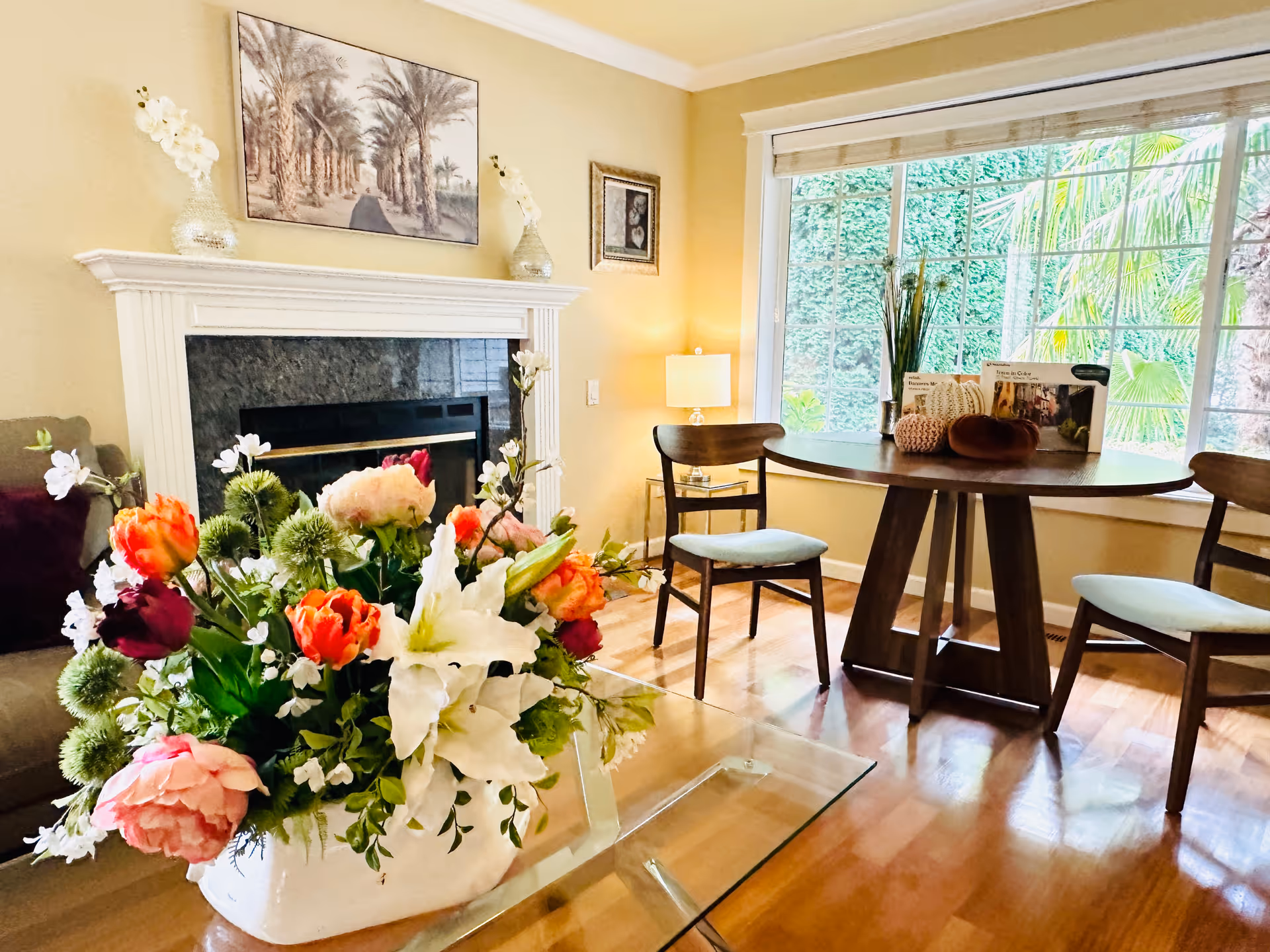 A cozy living room with a glass coffee table holding a large floral arrangement in the foreground. Behind it is a fireplace with a white mantel, decorated with vases and artwork above. To the right, there is a round wooden table with two chairs near a large window showing greenery outside. The room has warm lighting and wooden flooring.