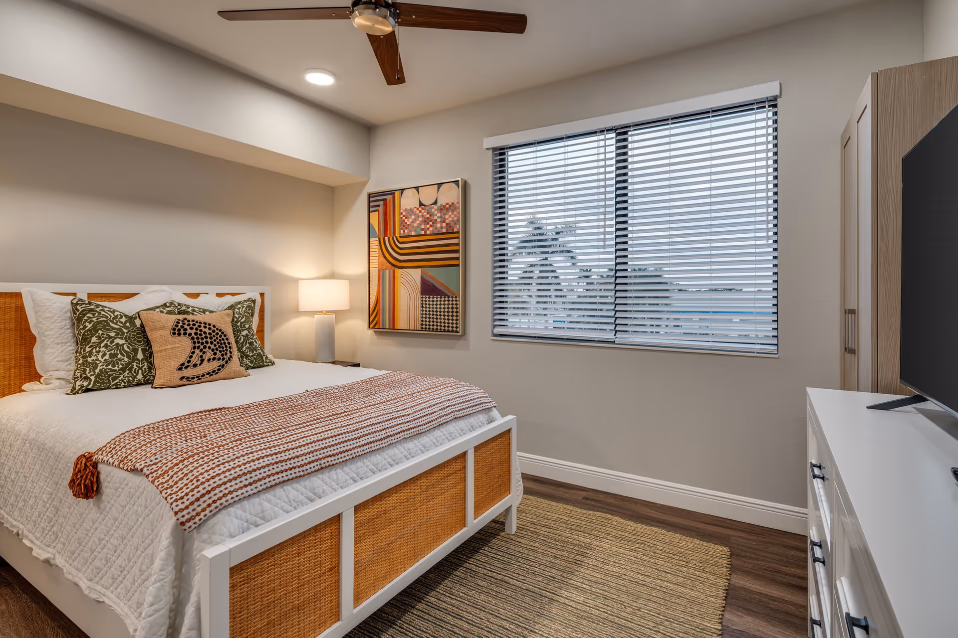 A cozy bedroom with a white bed frame featuring woven rattan panels, a white quilted bedspread, and decorative pillows including one with a bear design. There is a striped throw blanket at the foot of the bed. A modern table lamp sits on a nightstand next to the bed, and a colorful abstract painting hangs on the wall. The room has a window with horizontal blinds, a ceiling fan with wooden blades, a white dresser with a TV on top, and wood flooring with a woven rug.