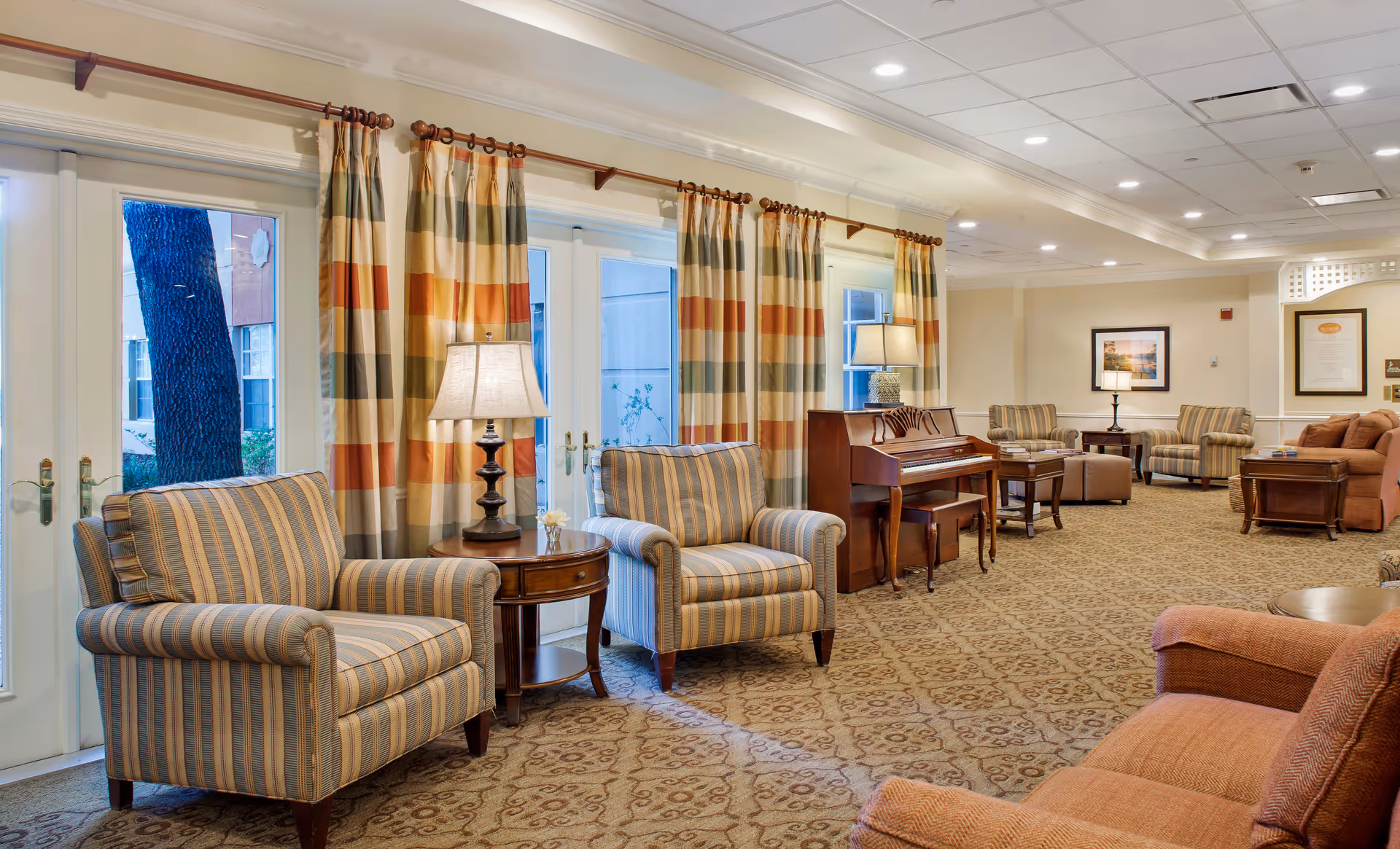 Bright, furnished senior living lounge with striped armchairs, sofas, side tables, lamps, a piano, and patterned carpet.