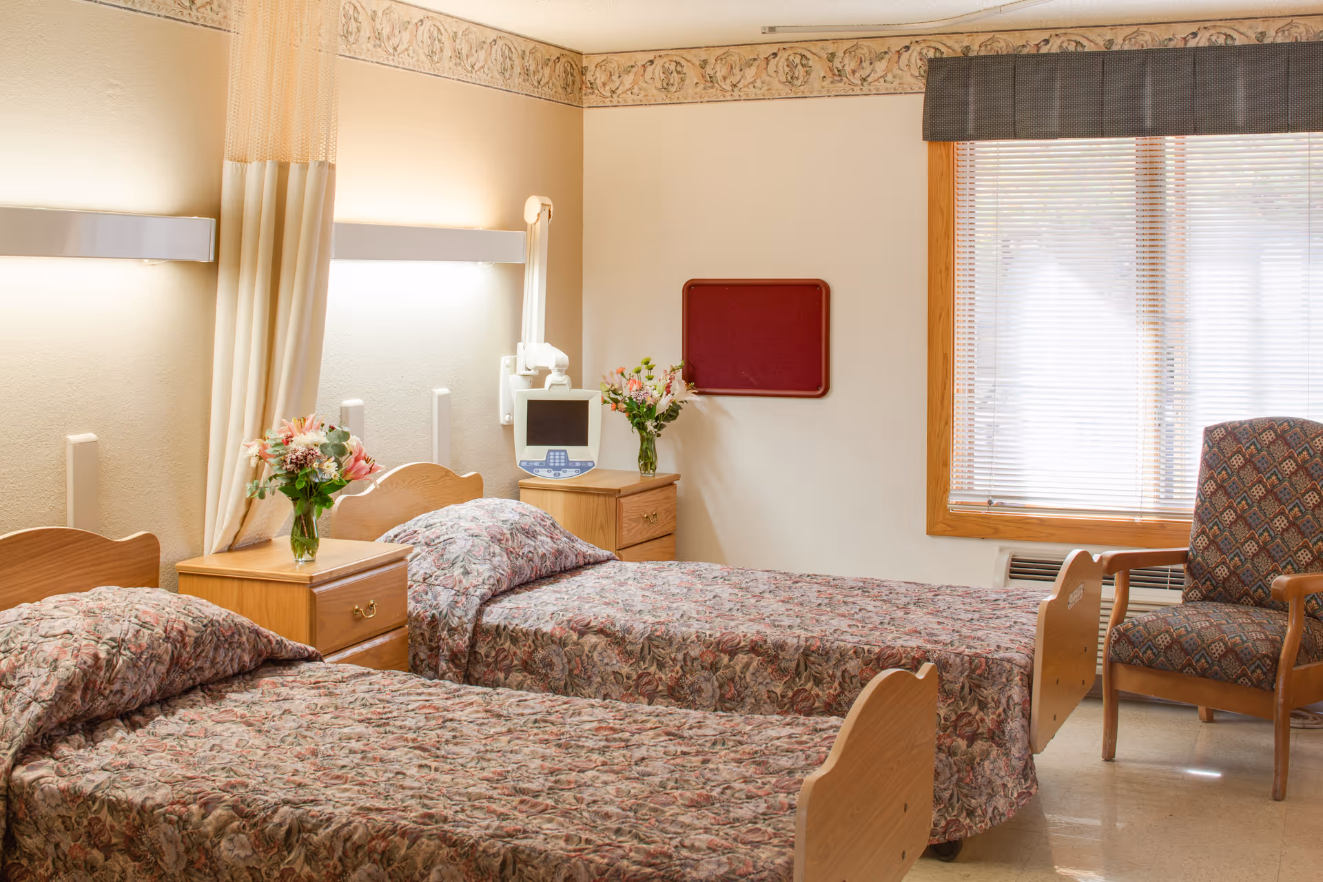 Two twin beds with floral bedspreads in a senior living bedroom, with nightstands holding vases of flowers, a window with blinds, and an upholstered armchair.