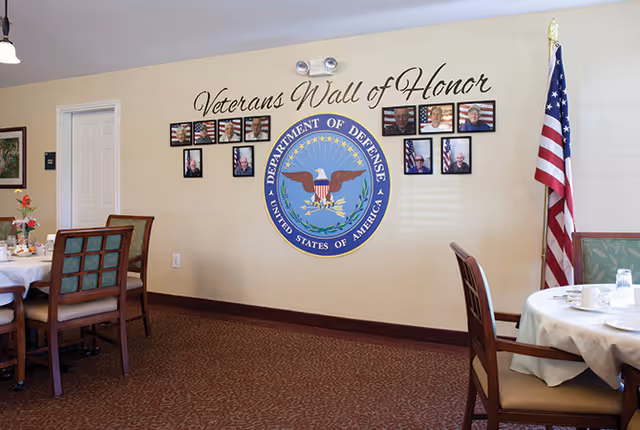 Dining room with round tables and chairs, an American flag, and a 'Veterans Wall of Honor' display featuring the Department of Defense seal on the wall.