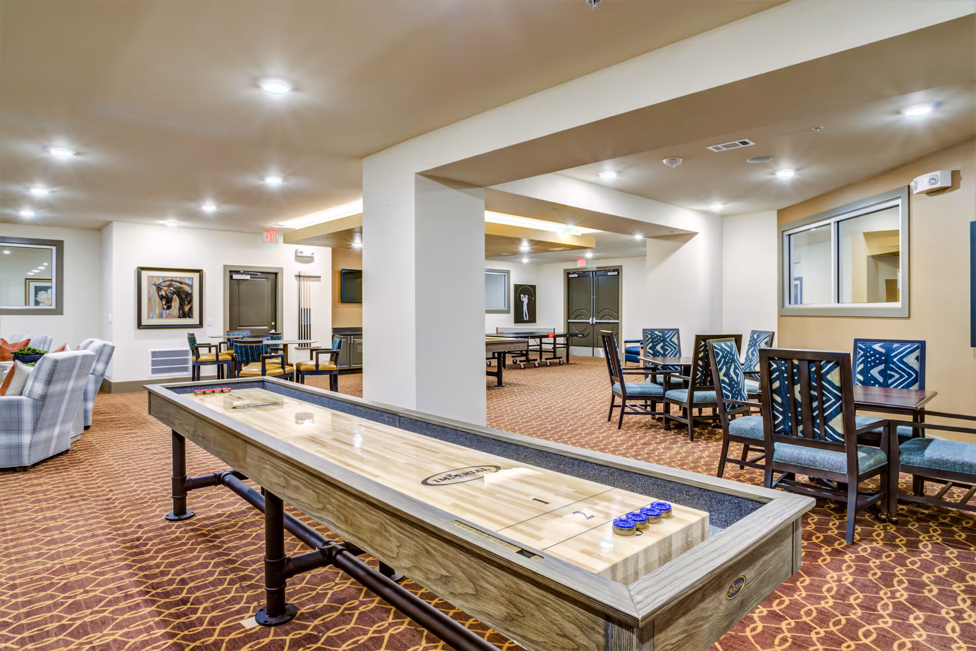 A spacious indoor recreational room with a shuffleboard table in the foreground, several tables and chairs with patterned upholstery, a ping pong table in the background, and framed artwork on the walls. The room has warm lighting and carpeted floors with a geometric pattern.