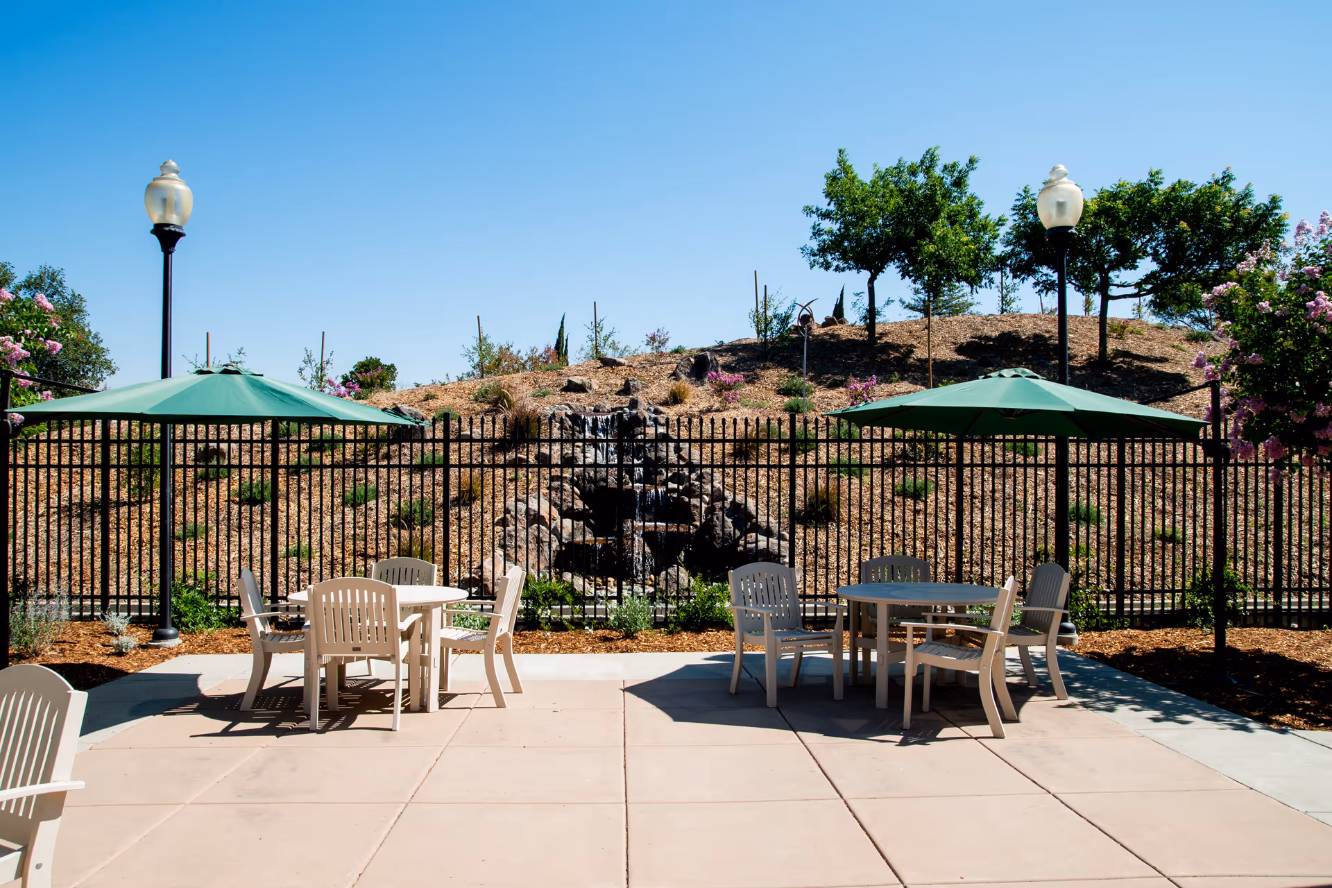 Outdoor patio area with two round tables, each surrounded by four white chairs and shaded by green umbrellas. Behind the patio is a black metal fence with a small waterfall feature and landscaped hillside with trees and flowering bushes under a clear blue sky.