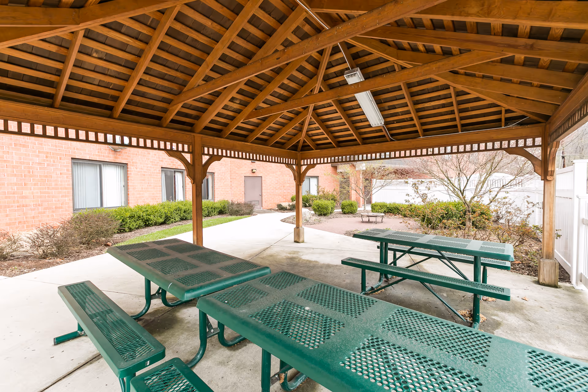 Outdoor covered pavilion with wooden roof structure and green metal picnic tables and benches on a concrete floor, surrounded by bushes and a brick building in the background.