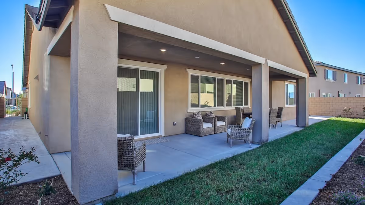 Covered patio area with wicker seating along the back of a beige assisted living building and a grassy strip.