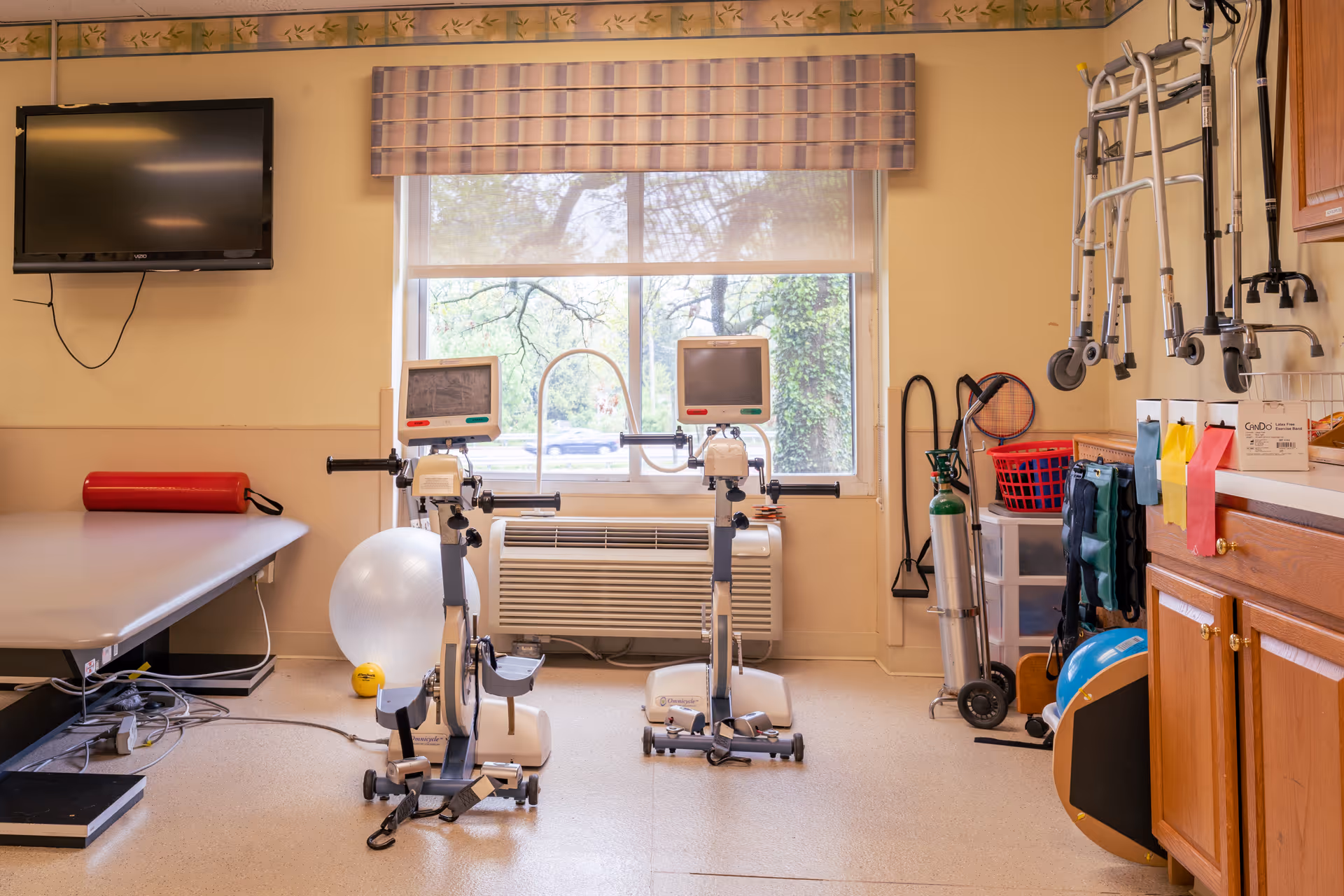 A therapy or rehabilitation room with exercise equipment including two stationary bikes with monitors, a large exercise ball, a red cylindrical bolster on a padded table, walkers hanging on the wall, an oxygen tank, and various therapy supplies on a countertop and shelves. A window with a striped valance lets in natural light.
