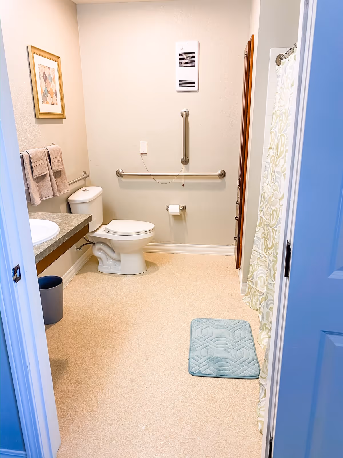 A clean and accessible bathroom with a toilet, a countertop with a sink, a trash bin, a towel rack with two towels, a framed picture on the wall, grab bars for support, a shower area with a patterned curtain, and a light blue bath mat on the floor.