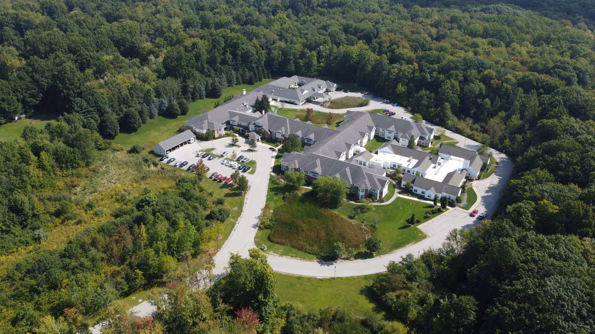 Aerial view of a senior living campus with multiple connected buildings, parking lots and a curved driveway surrounded by dense trees.